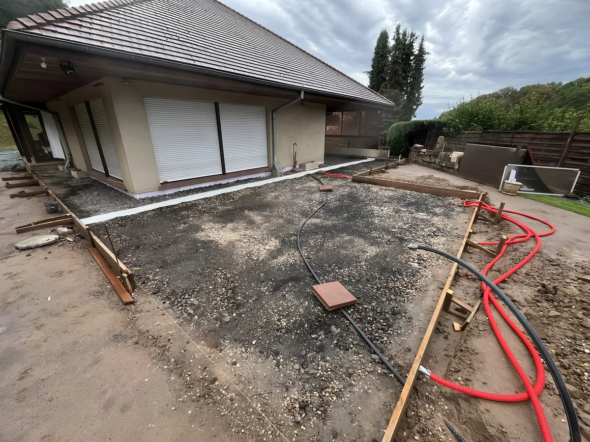 Coffrage bois et fond de forme en gravier préparés devant une maison avant le coulage d'une dalle béton, gaines en attente