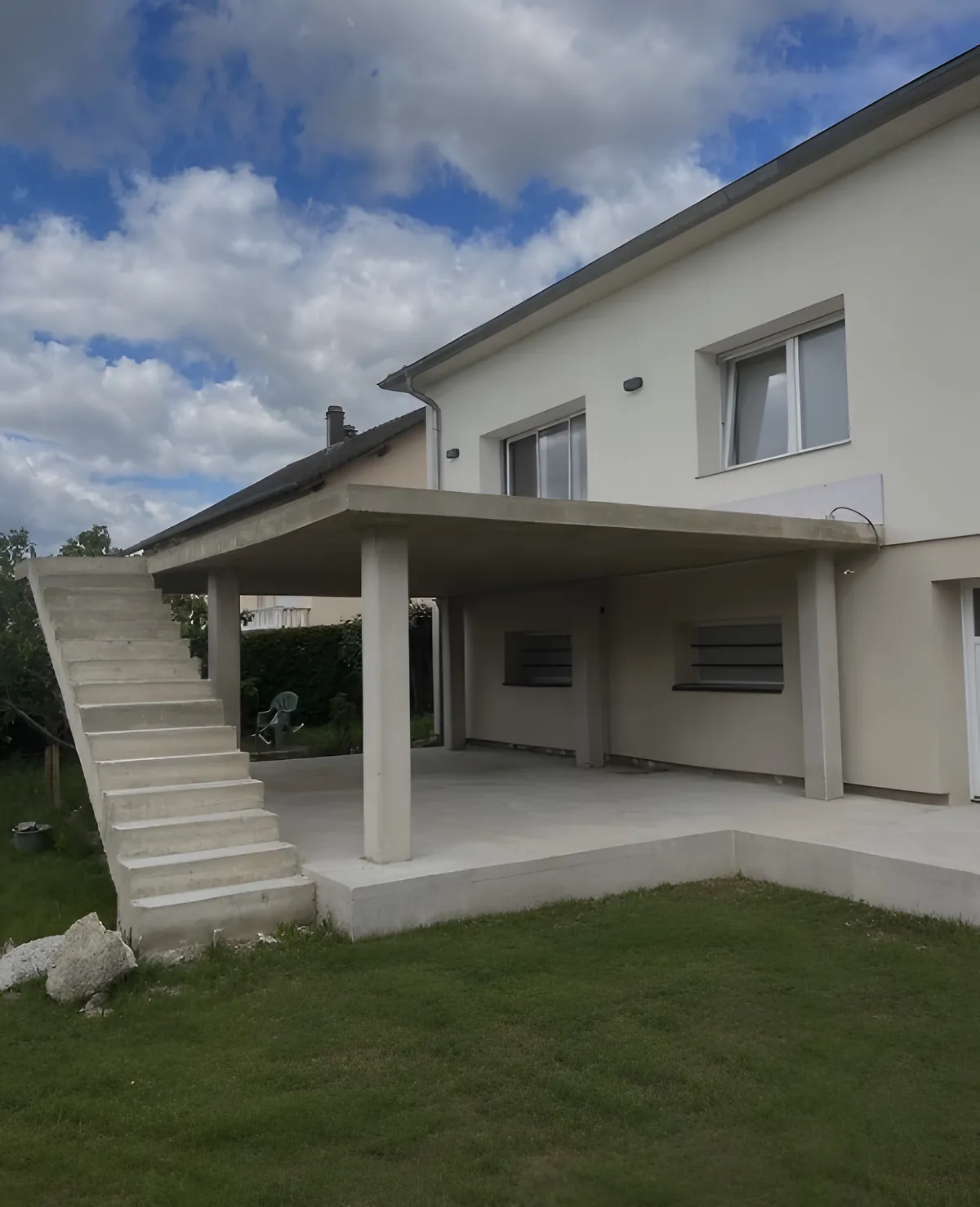 Terrasse couverte en béton sur poteaux, avec escalier extérieur en béton, à l'arrière d'une maison